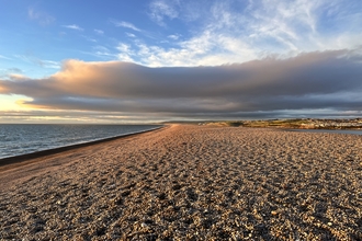 A view of Chesil Beach, a large bank of pebbles with the sea on one side and clouds above.