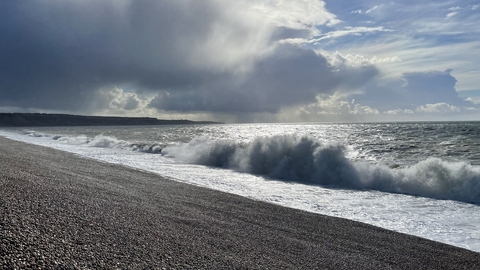 Big waves crashing up onto Chesil Beach, under cloudy, overcast skies.