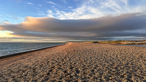 A view of Chesil Beach, a large bank of pebbles with the sea on one side and clouds above.