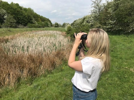 A teenage girl is looking for birds through binoculars, over a field surrounded by trees