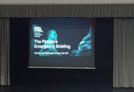 Cinema screen on a theatre stage, with an image of a man and the words The People's Emergency Briefing