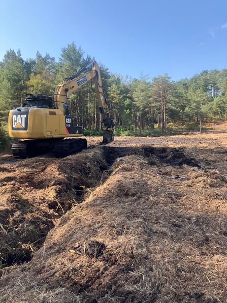 A digger shown filling in a deep ditch with a forest in the background
