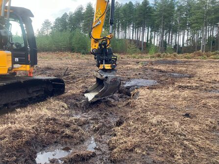 A digger smoothing ridges and furrows in a wet peat landscape with trees in the background