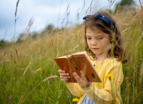 A young girl reading an old wildlife identification book sat amongst long green grass